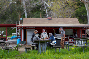 A group of people eating at a picnic table in front of a red cabin.